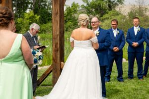 Outdoor wedding ceremony under wooden pergola with wedding party in blue suits and mint green dress.