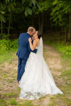 Wedding couple embracing on forest path with bride in flowing white gown and groom in navy suit.