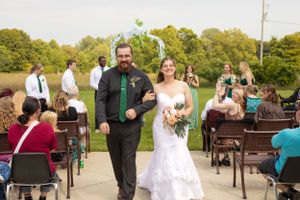 Newlyweds walk down the aisle at an outdoor ceremony while guests look on from wooden chairs.