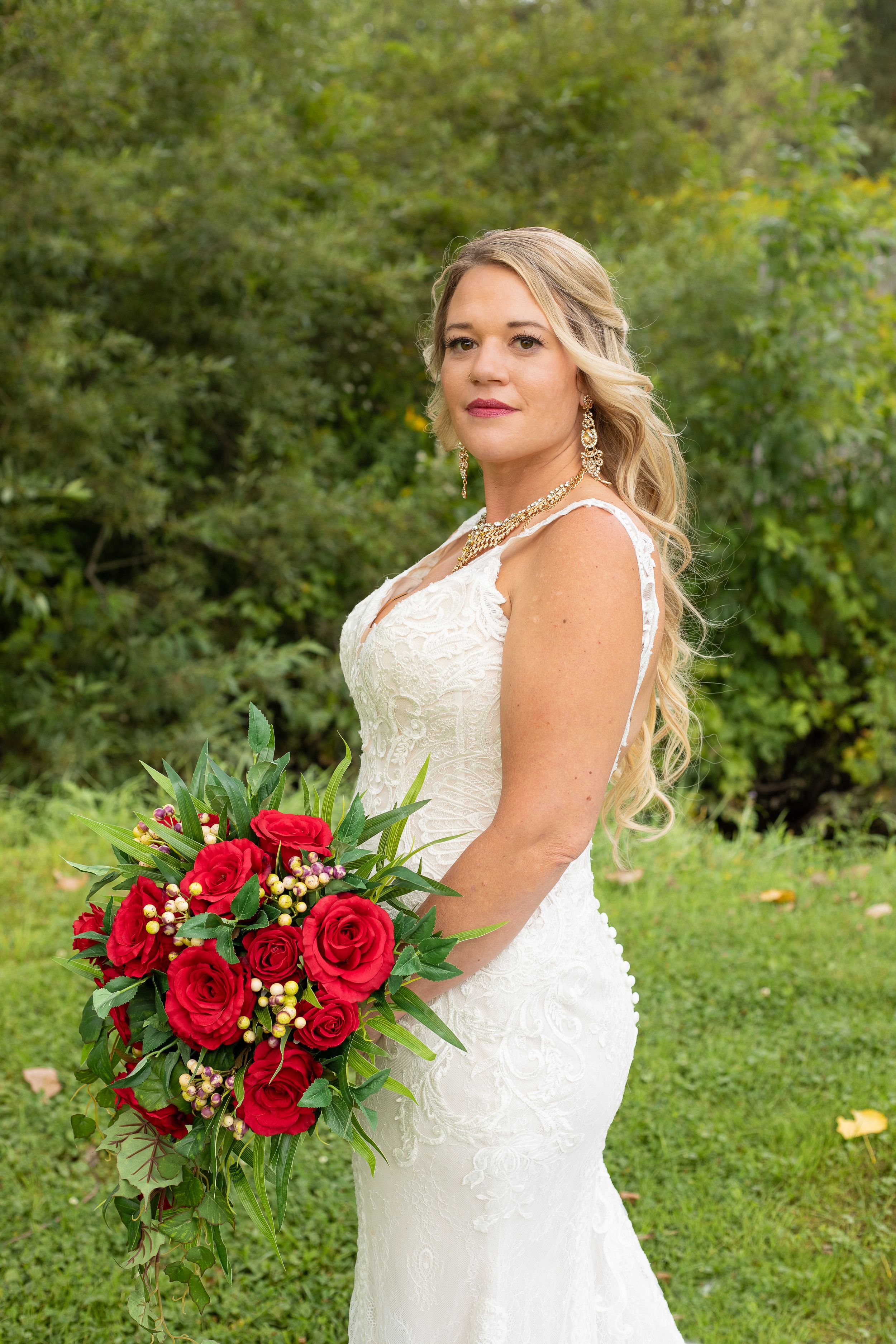 Elegant bride in white dress holds vibrant red rose bouquet against lush garden backdrop.