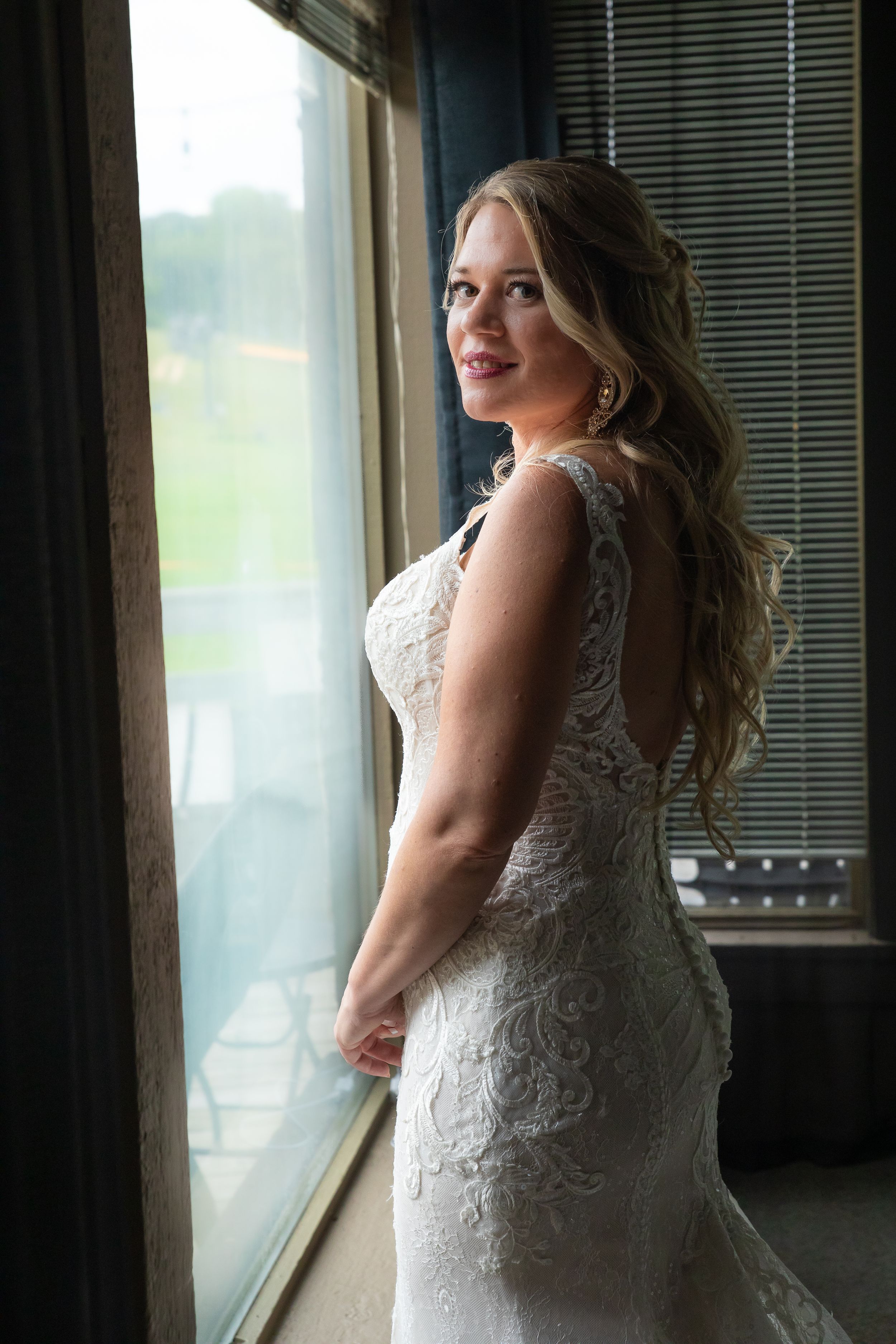 A bride in lace dress poses by window with natural light highlighting her silhouette.