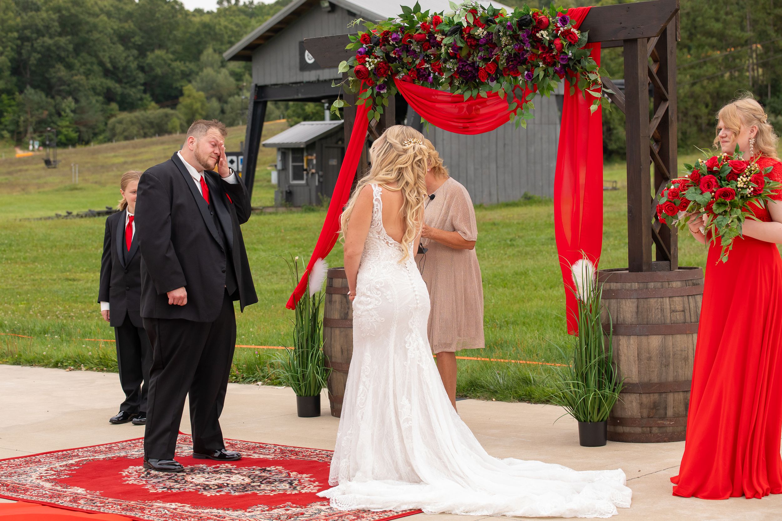 Wedding ceremony setup features red floral arch and red carpet in rustic outdoor setting.