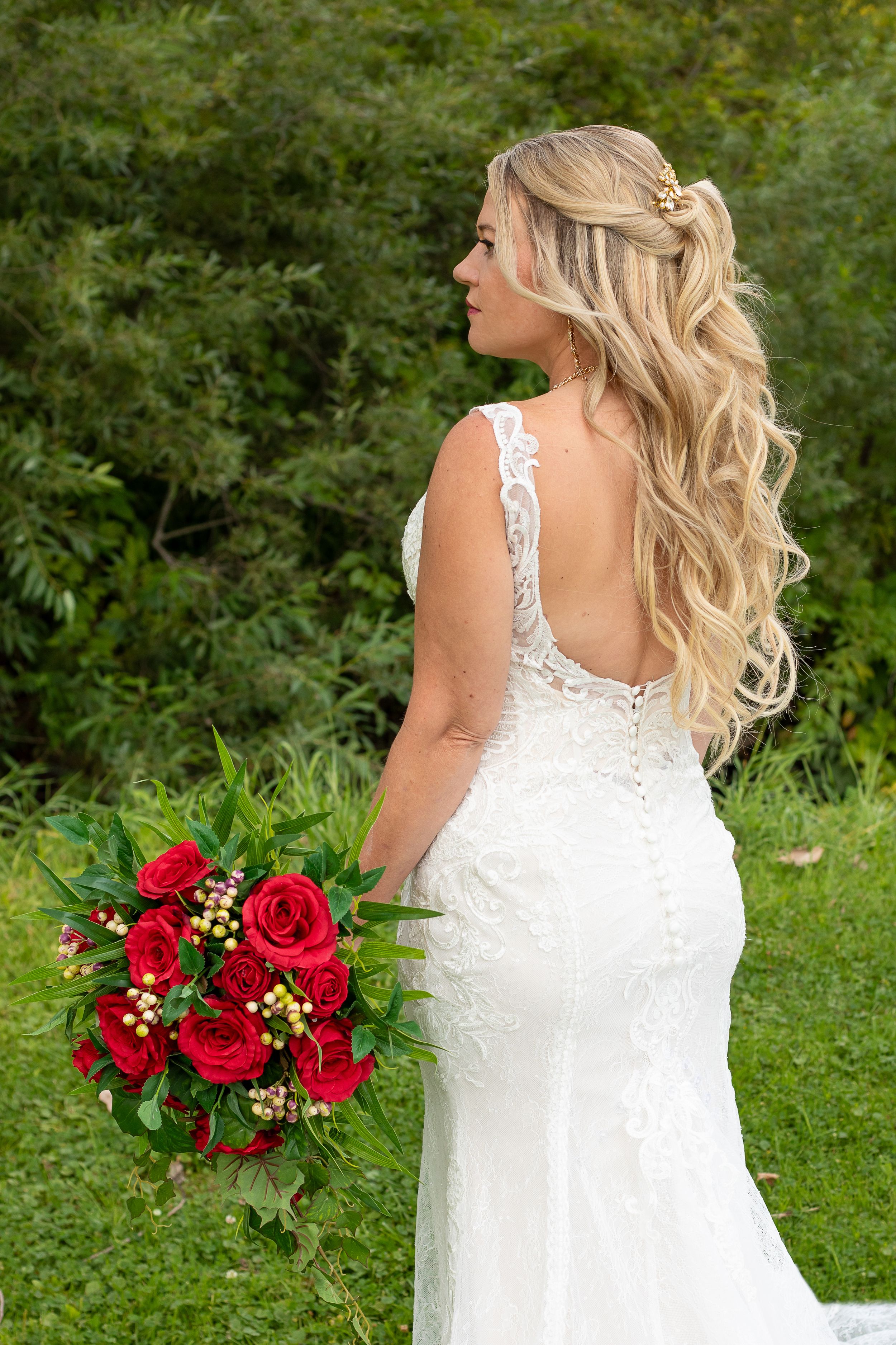 Back view of a bride in a lace wedding gown holding red roses outdoors.