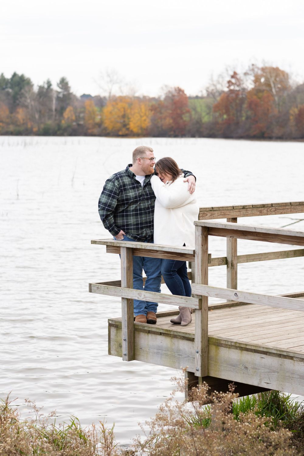 Couple sharing a moment on wooden dock overlooking lake with fall foliage in background.