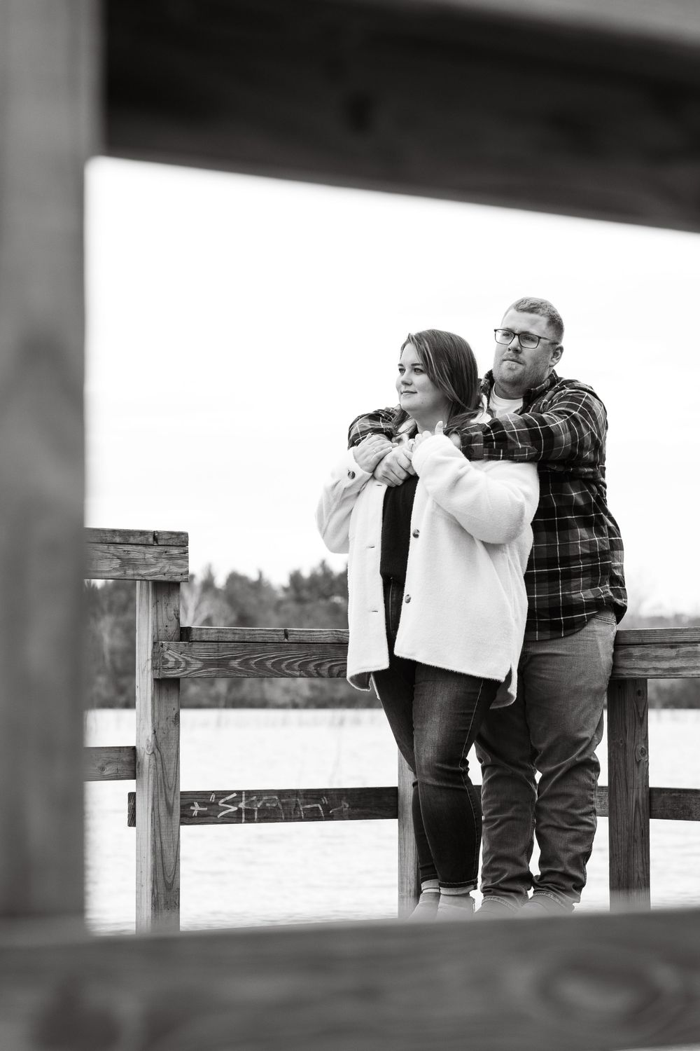 Black and white photo of couple embracing on wooden dock over water.
