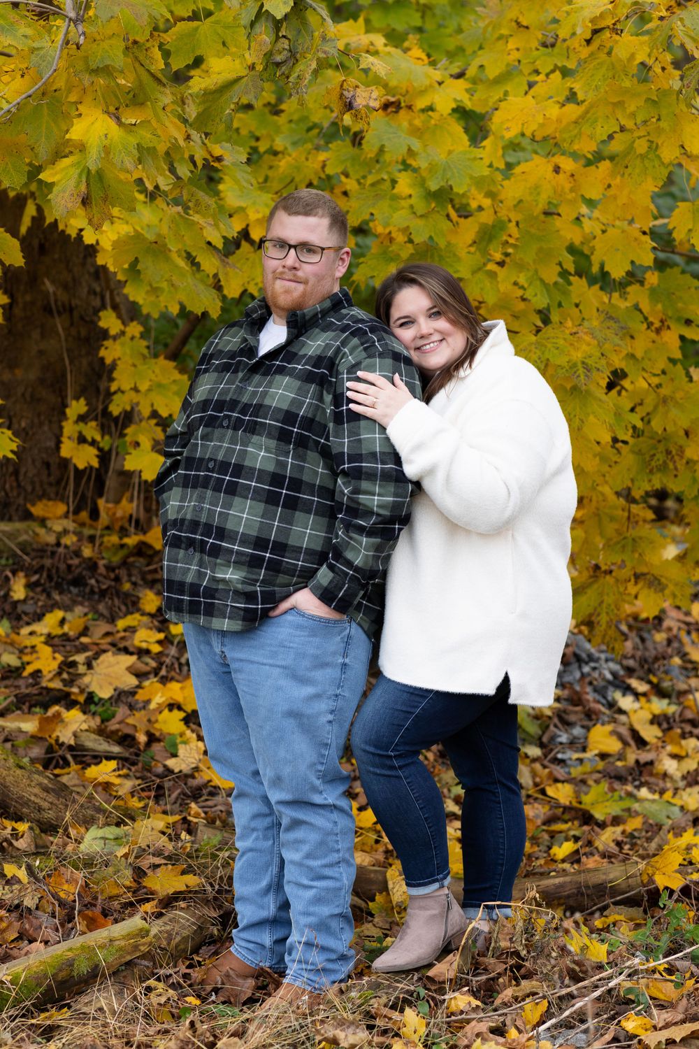 A couple poses together in front of vibrant yellow autumn leaves wearing plaid and white sweaters.