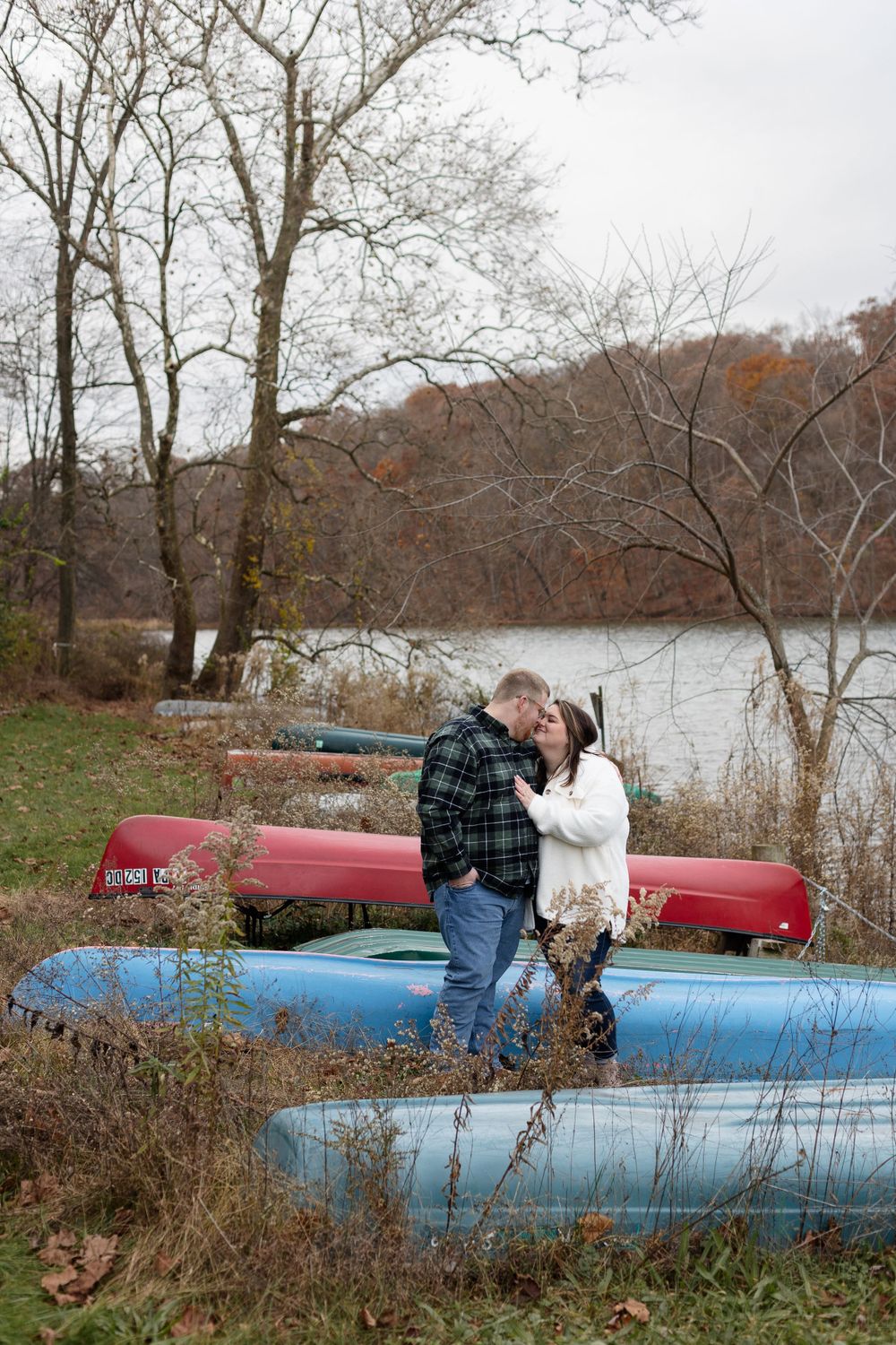 A couple stands together on stacked kayaks by a lake during autumn.
