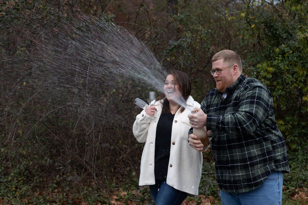 A couple sprays champagne outdoors while celebrating in casual fall attire.