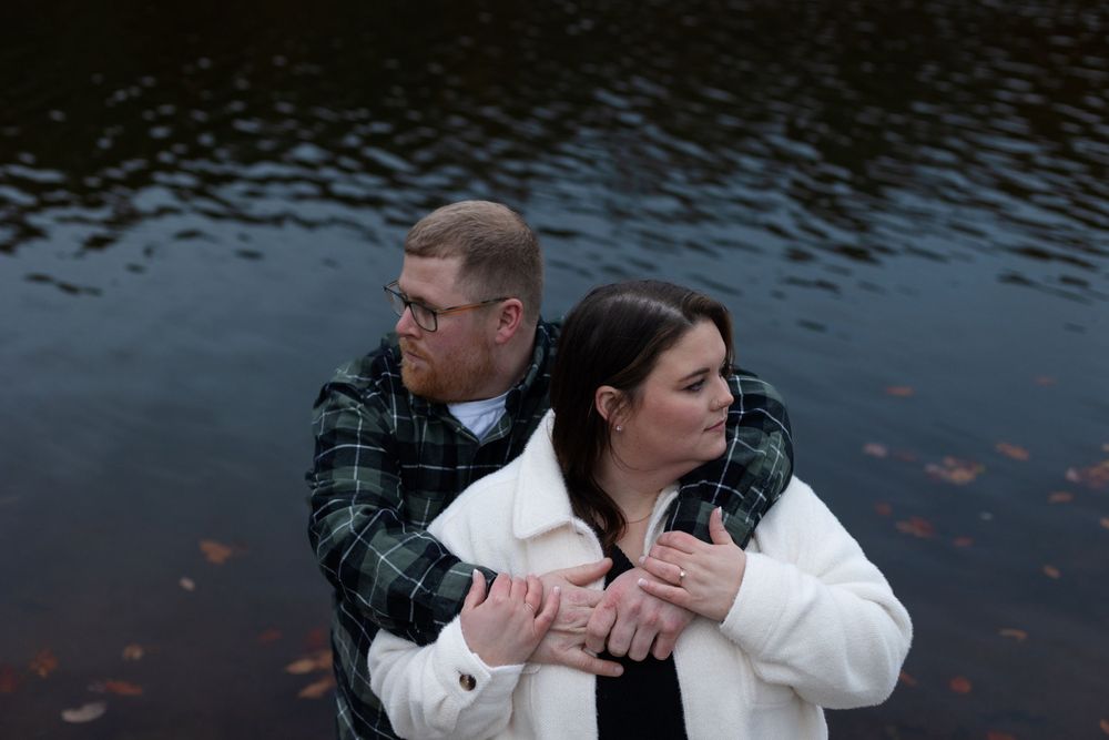 A couple embraces by a lake with autumn leaves floating on the water's surface.