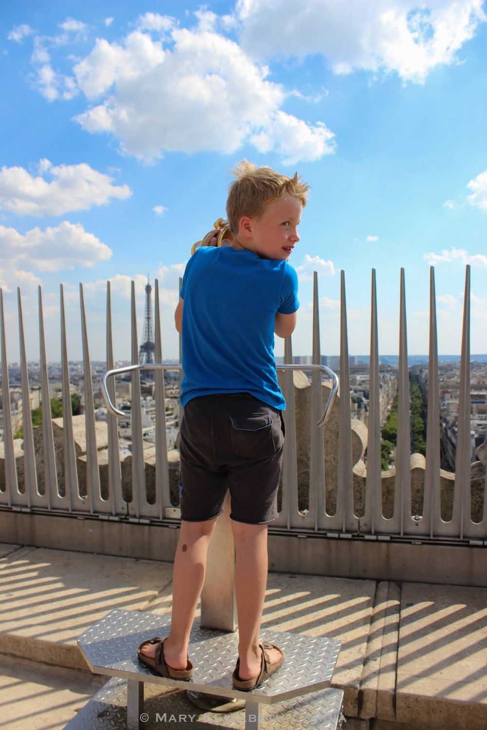 Child in blue shirt looking through observation deck railing at city view below.
