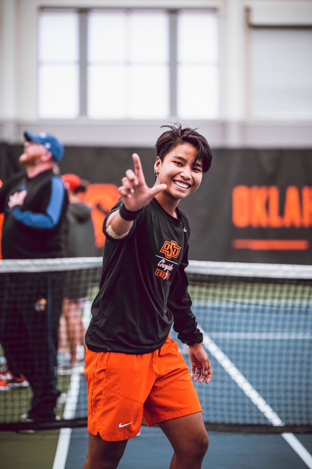A tennis player in Oklahoma State gear gestures towards the camera on an indoor court.