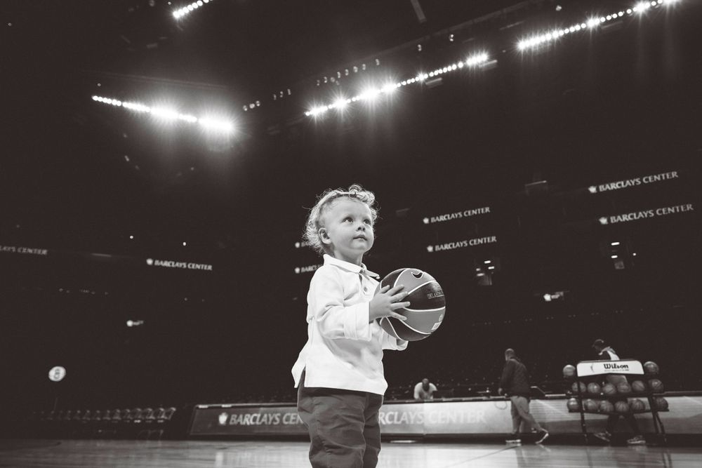 A small child holds a basketball on a dimly lit court with stadium lights in the background.