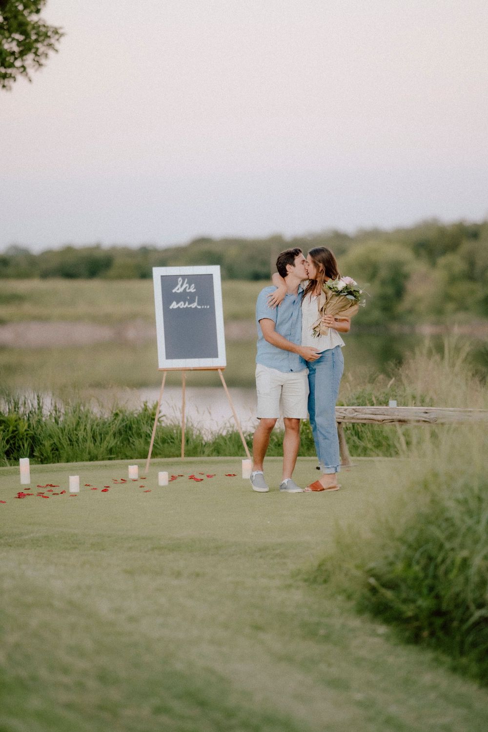 A couple shares a romantic moment outdoors next to a chalkboard sign during a surprise proposal photoshoot.