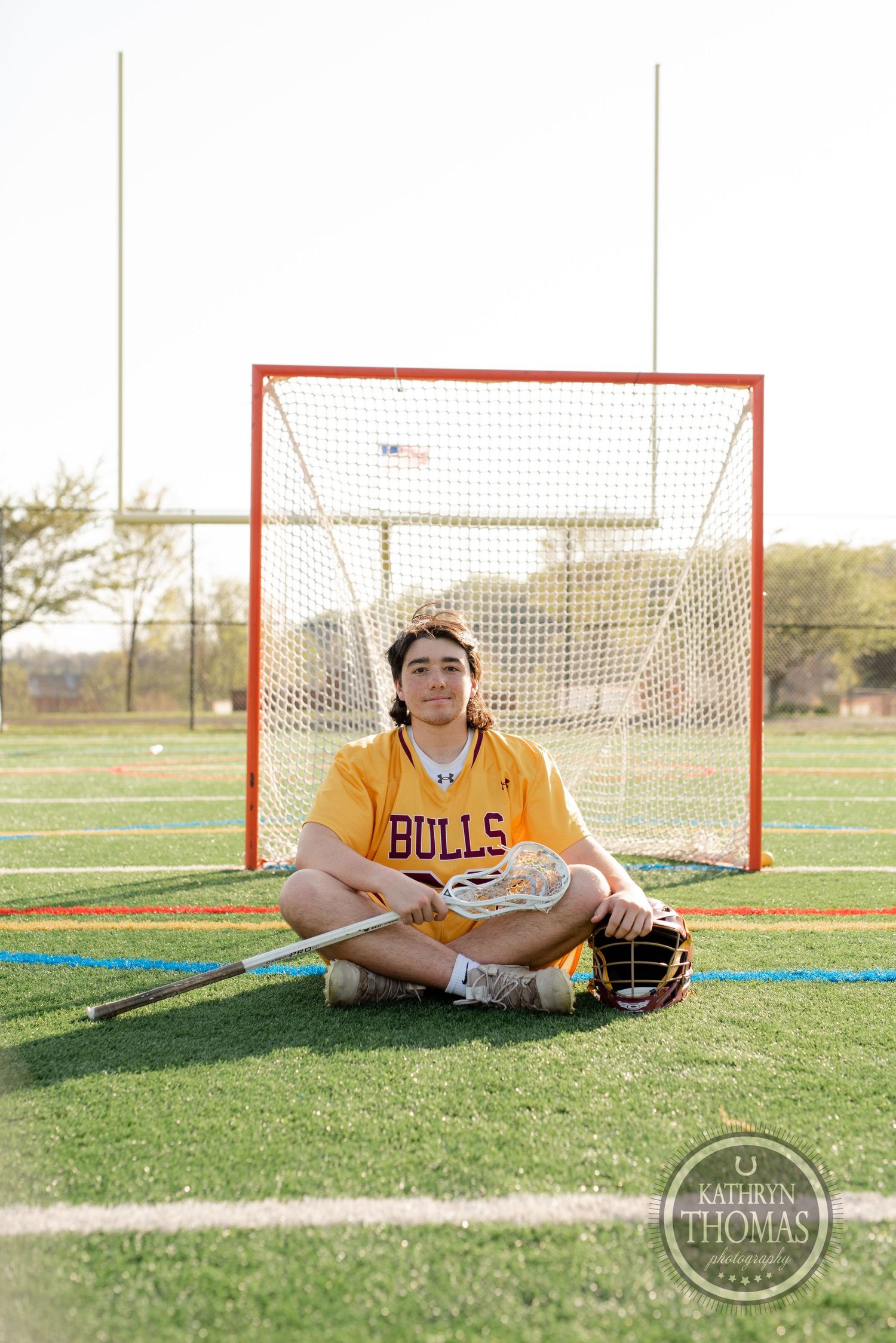 Lacrosse player in yellow Bulls jersey sitting with equipment in front of goal net.