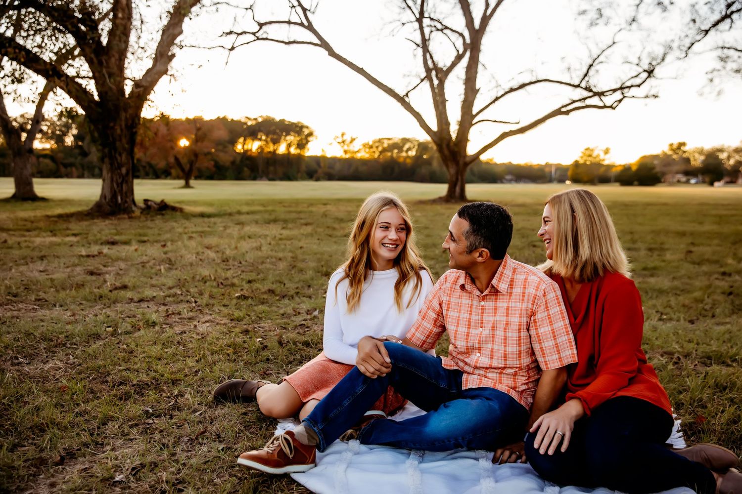 Three friends sitting together on a blanket during sunset in an open field.