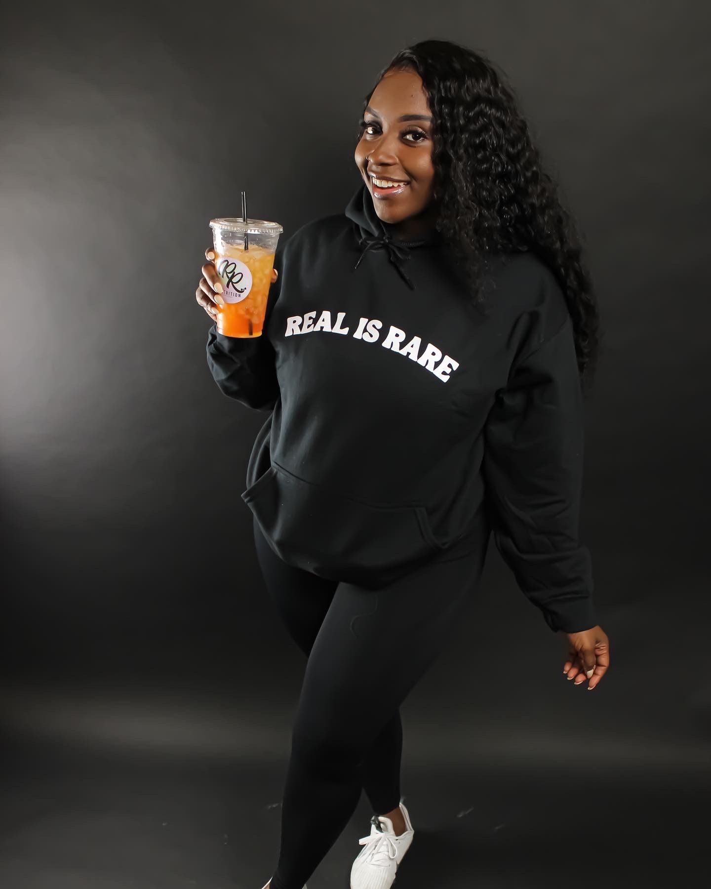 A person in a black 'Real is Rare' sweatshirt holds an iced drink while posing against a dark background.
