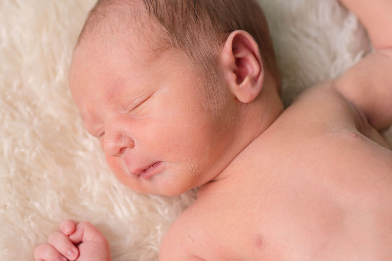 Newborn baby laying on pillow. He looks to have a smile and wink while he is about to chew his fist.