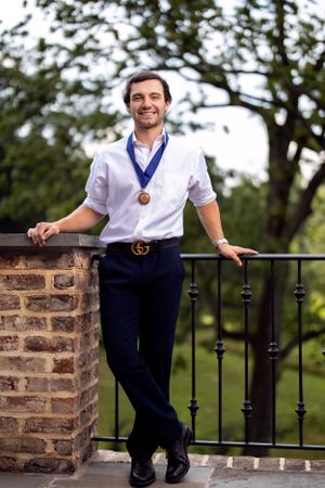 Graduate wearing medal and white shirt poses by stone wall railing in outdoor setting.