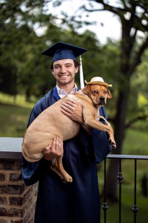 Graduate in academic regalia holding brown dog while posing outdoors.