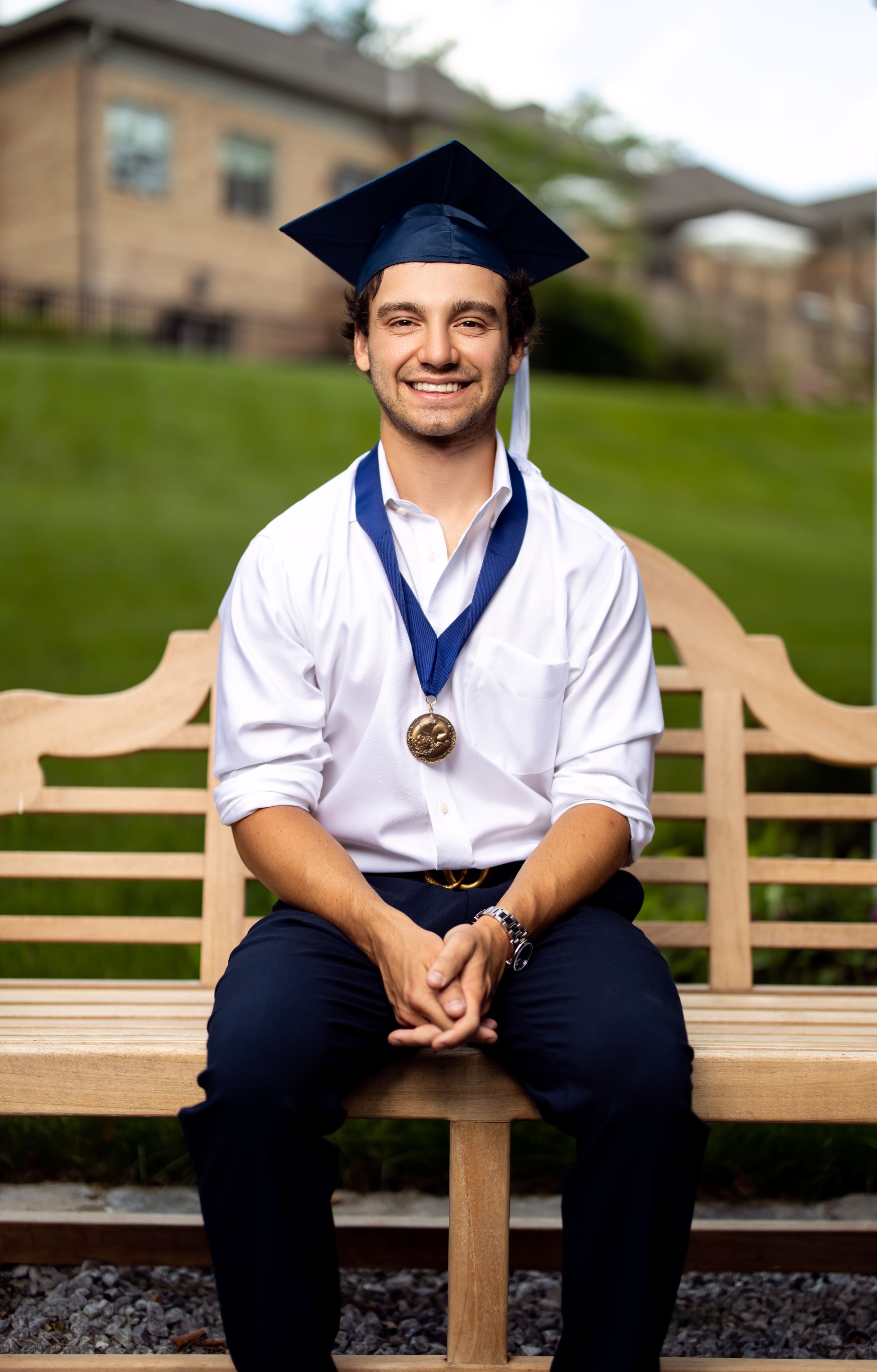 Graduate in cap and gown sits on wooden bench outdoors with medal around neck.