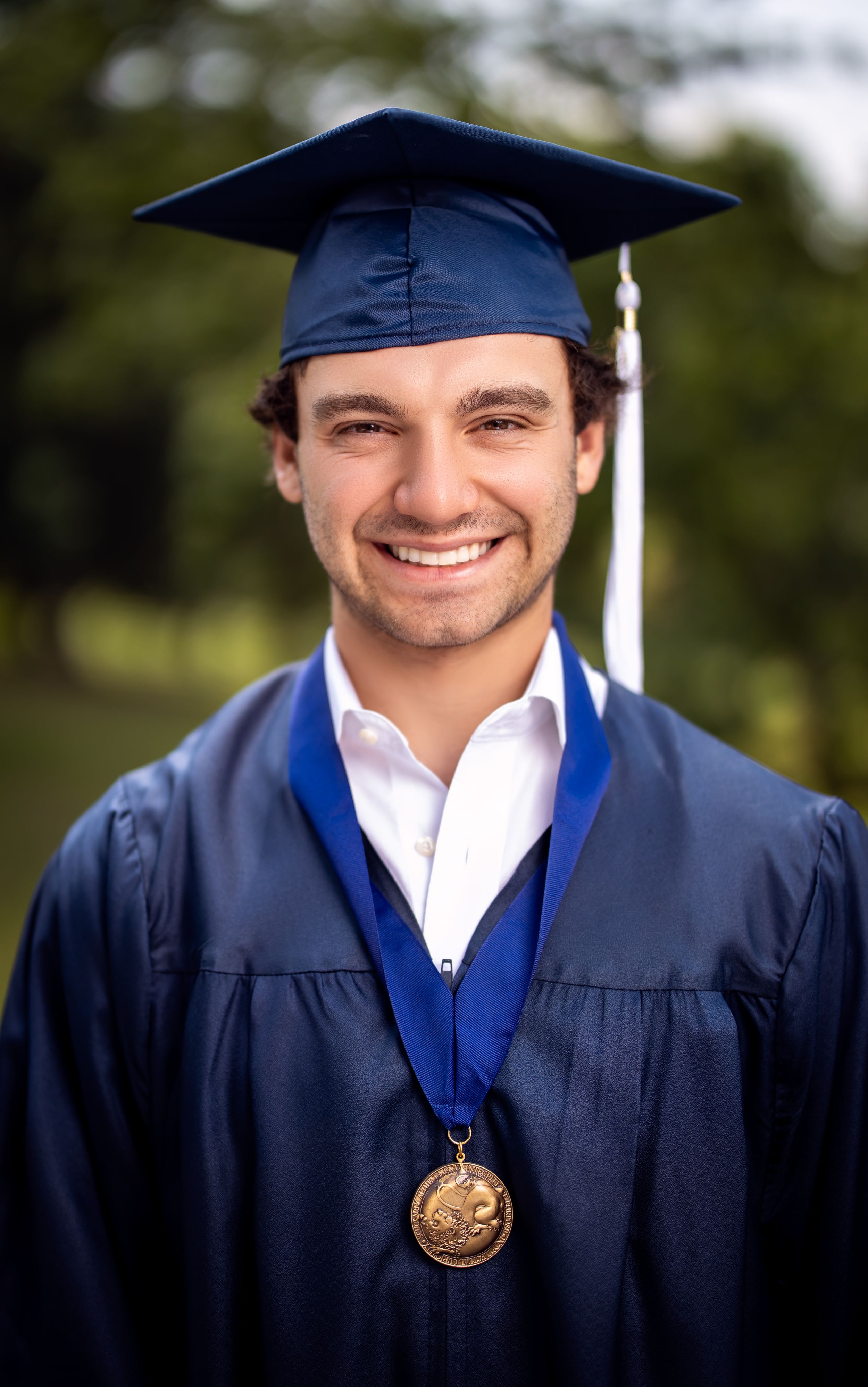 Graduate in navy blue cap and gown wearing medal against blurred outdoor background.