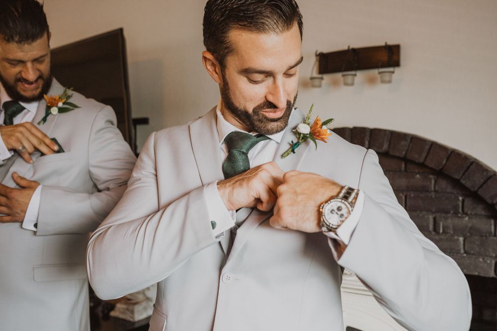 Groomsmen in light suits help each other with boutonnieres during wedding preparations.