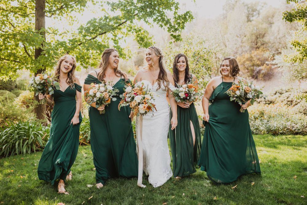 Wedding party in emerald green dresses laughing and dancing together in a sunny outdoor garden setting.