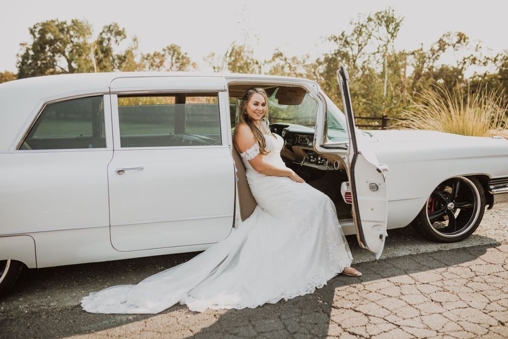 Bride in flowing white gown poses with vintage limousine during sunset photo session.
