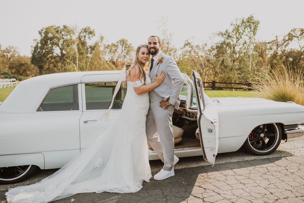 Wedding couple poses beside a pristine white vintage car in an outdoor setting.