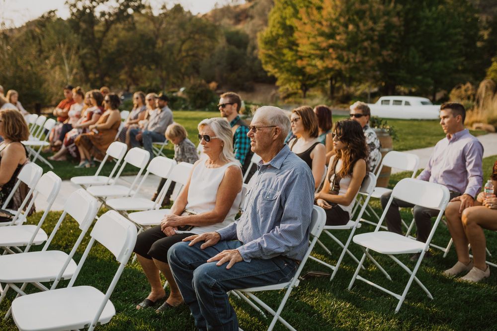 Wedding guests seated in white folding chairs on a lawn during an outdoor ceremony.