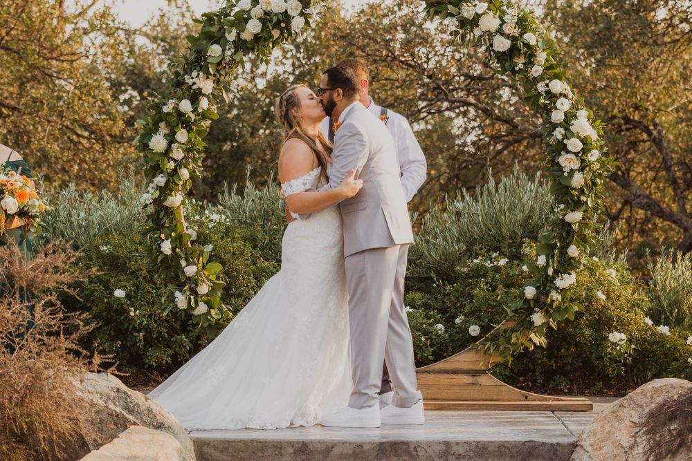 Couple embracing during sunset wedding ceremony with floral arch and natural backdrop.