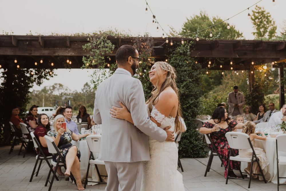 A romantic outdoor wedding dance scene with string lights and table settings in the background.