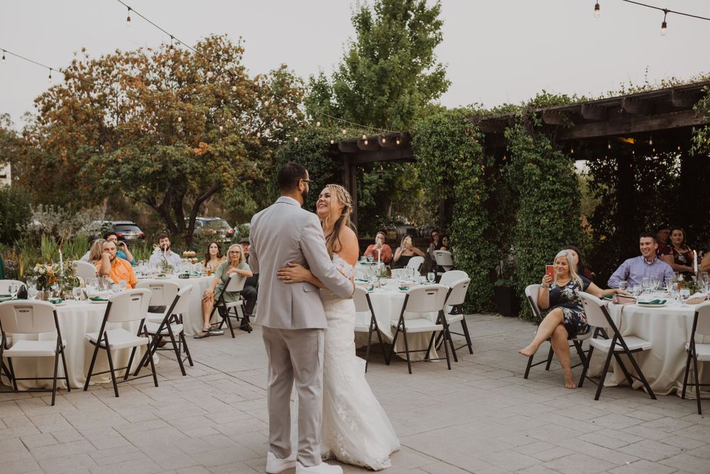 A couple shares their first dance on an outdoor patio surrounded by wedding reception tables and string lights.