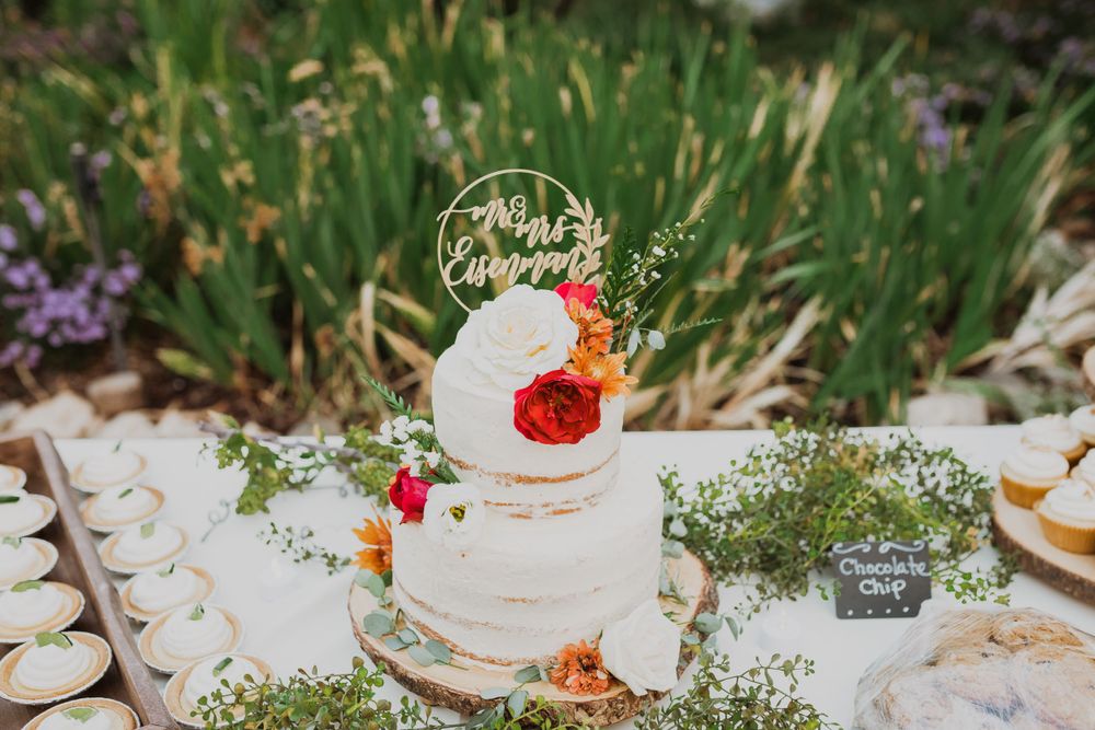 A white wedding cake decorated with fresh flowers and greenery sits on a table in a garden setting.