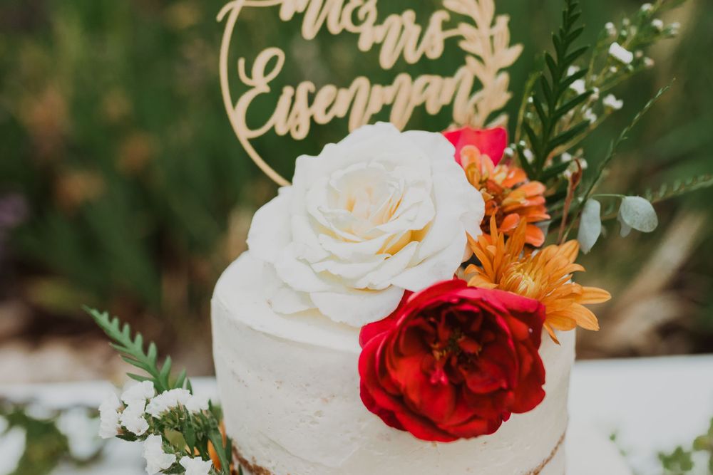 A white wedding cake topped with red and white roses and a gold cake topper against a garden background.