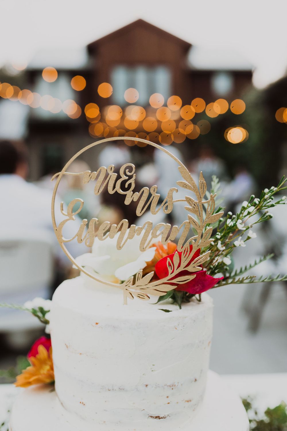 A white wedding cake with a gold circular topper and floral decorations against bokeh lights at dusk.