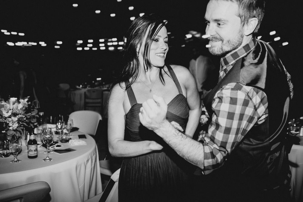 A black and white photo of guests dancing at an indoor wedding reception with tables in background.