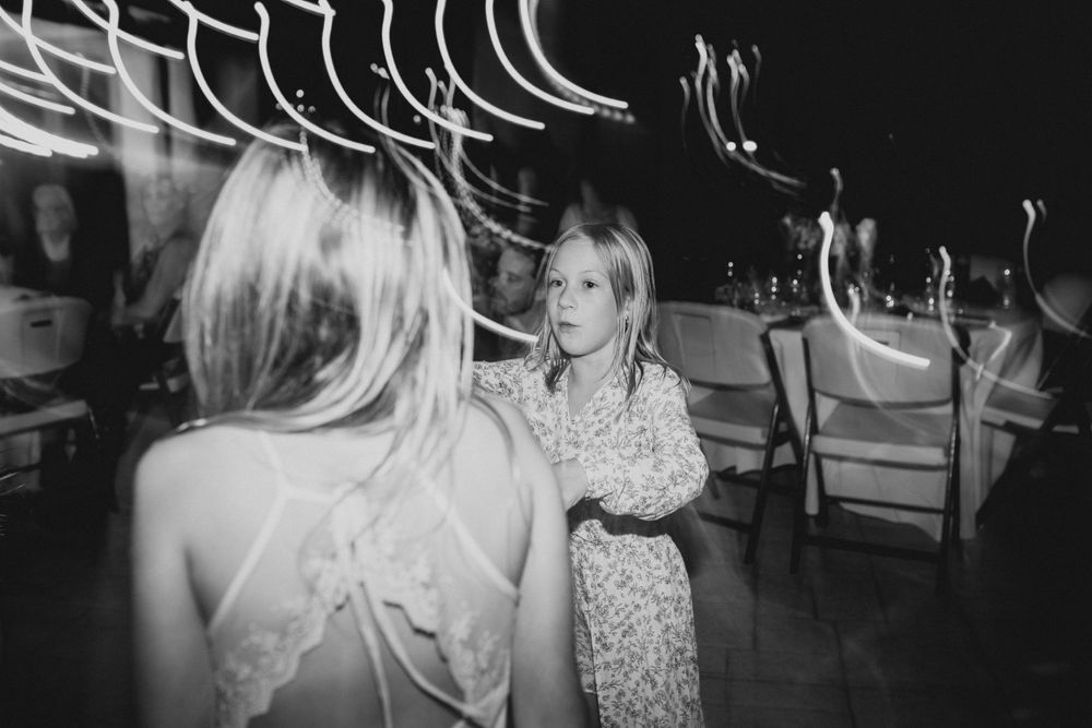 A black and white photo of guests dancing at a wedding reception with string lights overhead.