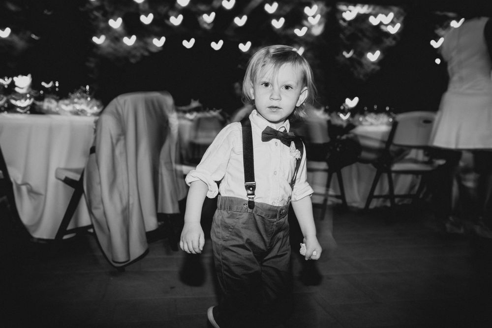 A young ring bearer in suspenders and bow tie stands under heart-shaped lights at a wedding reception.