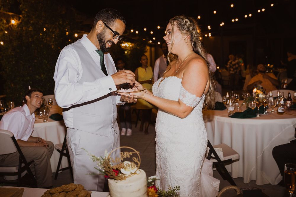 Newlyweds feeding each other cake during their wedding reception under twinkling lights.