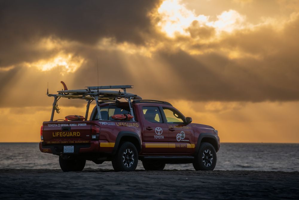 Red lifeguard pickup truck parked on beach at dramatic golden sunset with rays breaking through clouds.