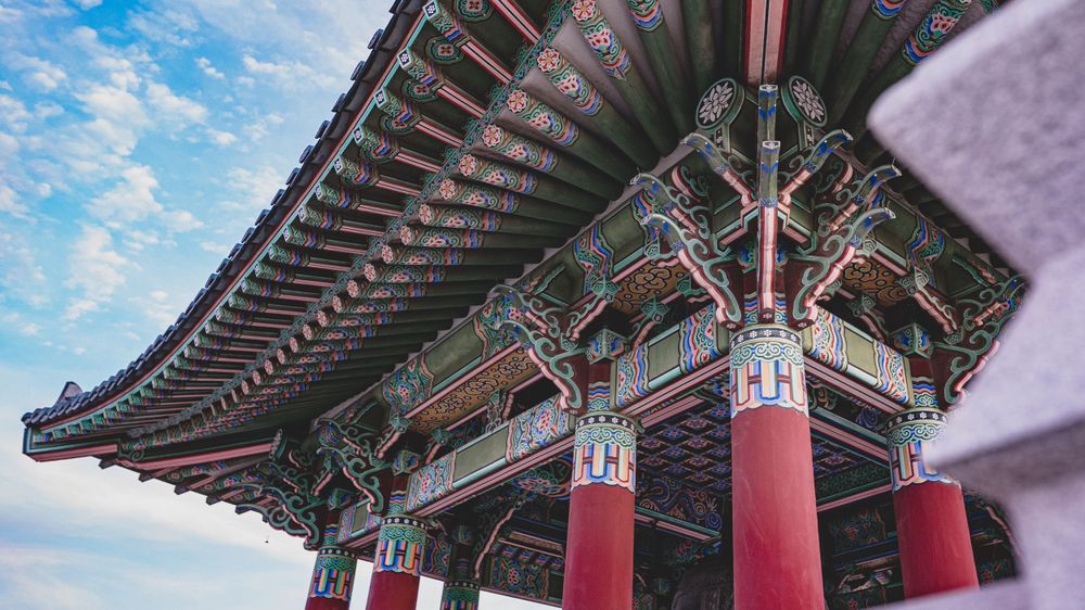 Ornate Korean traditional pavilion with colorful painted details, red columns, and curved tiled roof against a blue sky.
