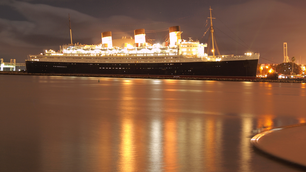 Historic RMS Queen Mary ocean liner illuminated at night reflecting golden lights on calm harbor water.