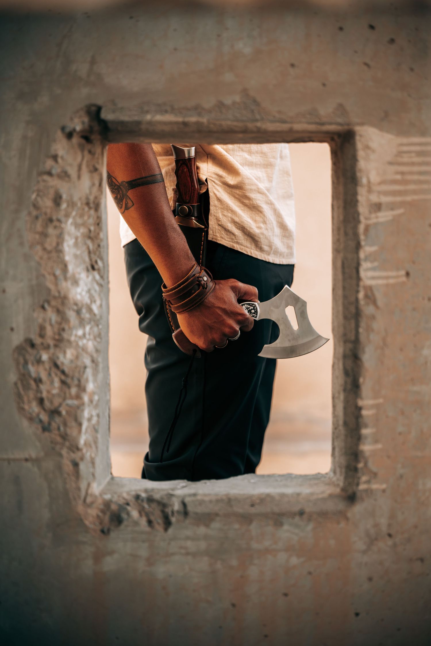 Tattooed person holding a small axe viewed through a square concrete wall opening.