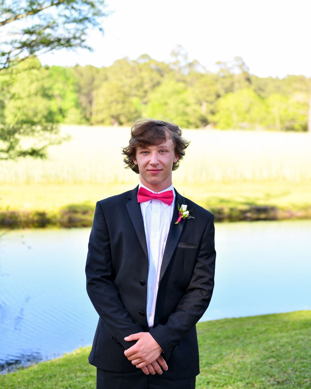 A person in formal wear with red bow tie poses outside by a pond with trees and grass in background.