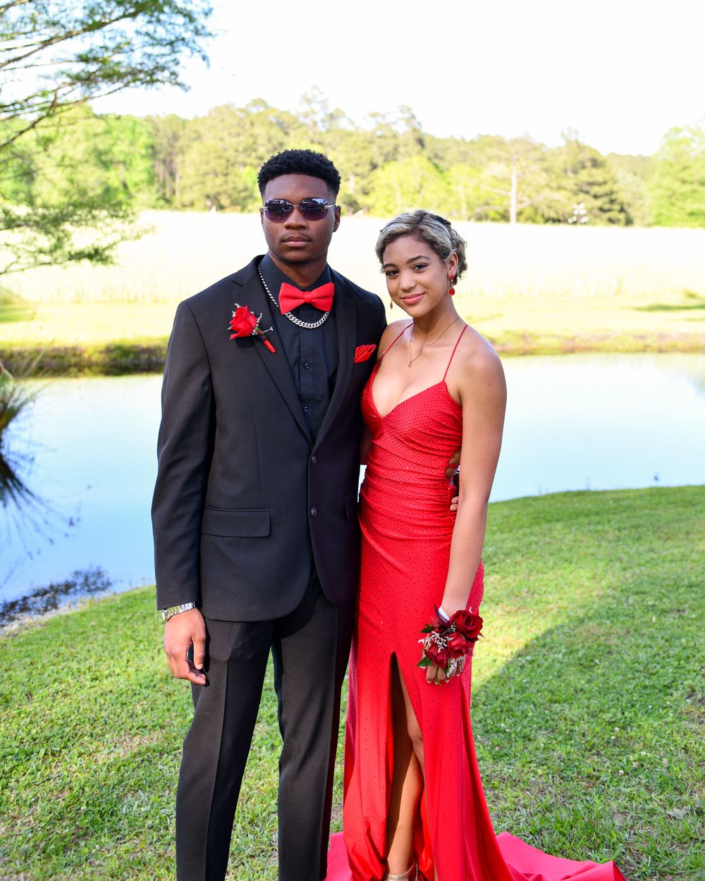A couple in formal attire poses for prom photos, with her in red and him in black by a scenic lake.