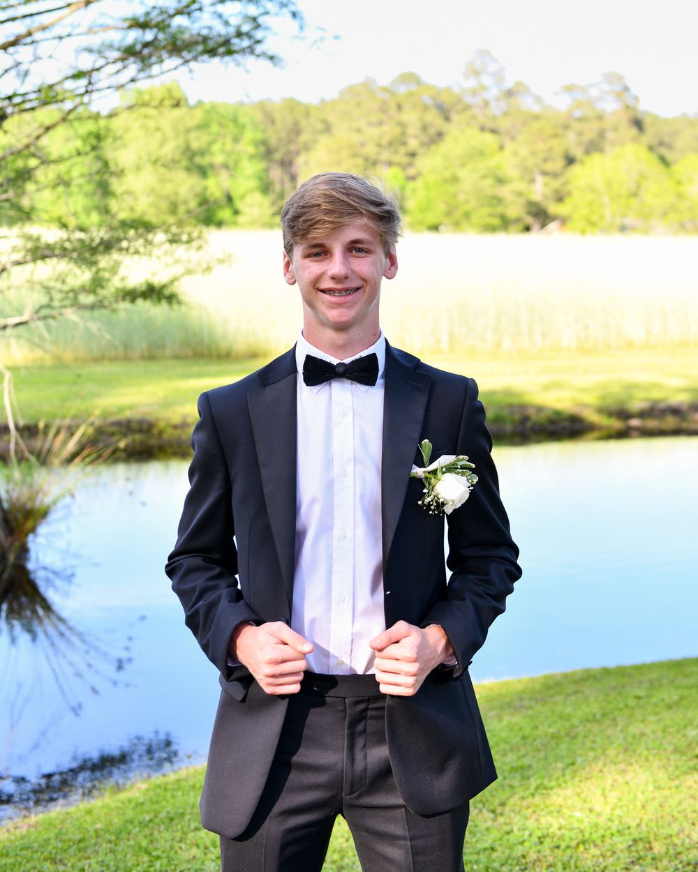 A person in a black tuxedo with bow tie stands by a lake on a sunny day holding their jacket.
