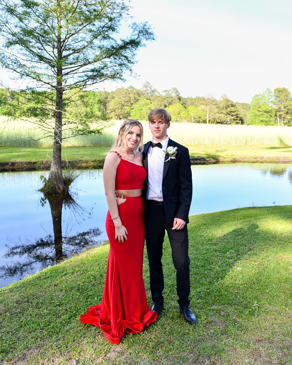 Couple in formal attire pose by lake, wearing red gown and black tuxedo.