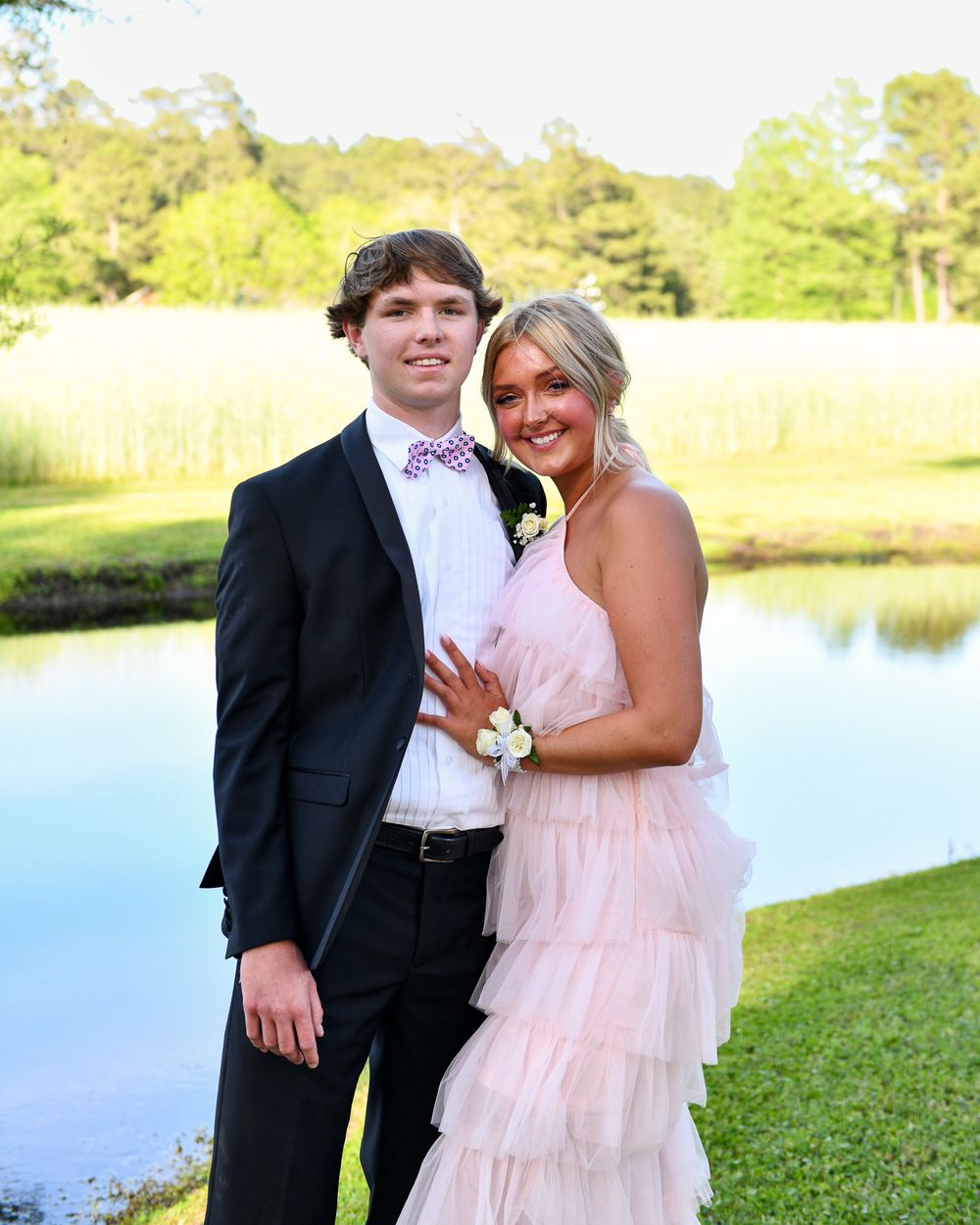 Formal couple poses lakeside with person wearing black tux and pink ruffle dress.