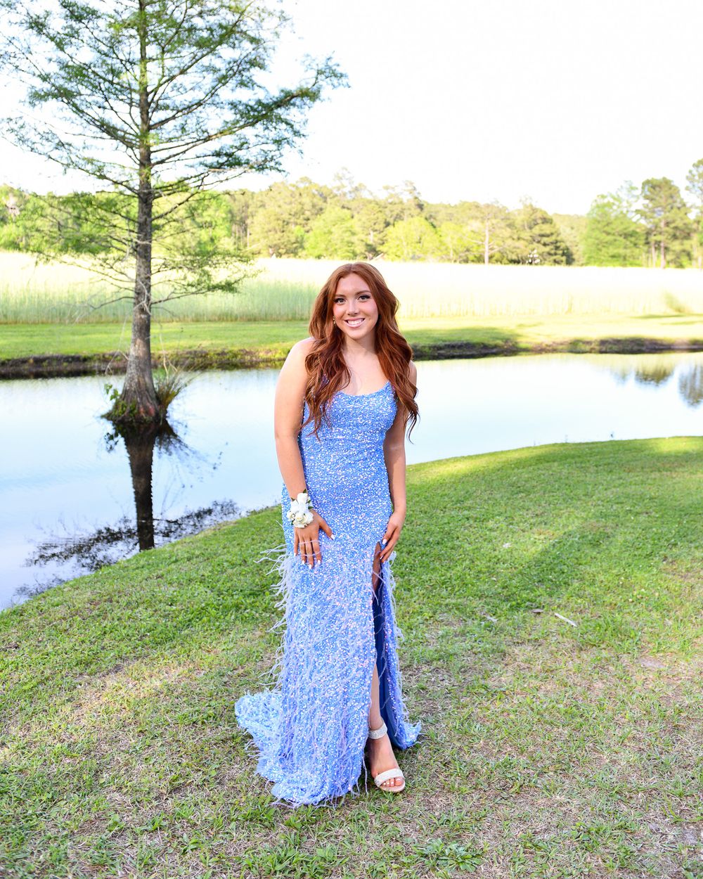 Woman in sparkly blue formal gown poses by lake with cypress tree backdrop.